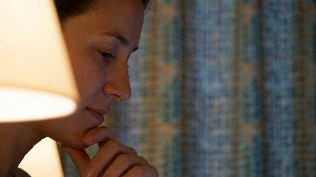 Femme vue de profil, le menton appuyé sur la main, regard baissé et air pensif, dans une pièce faiblement éclairée.