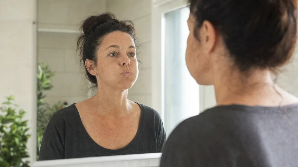 Femme réalisant un exercice de gymnastique faciale devant un miroir, les joues gonflées