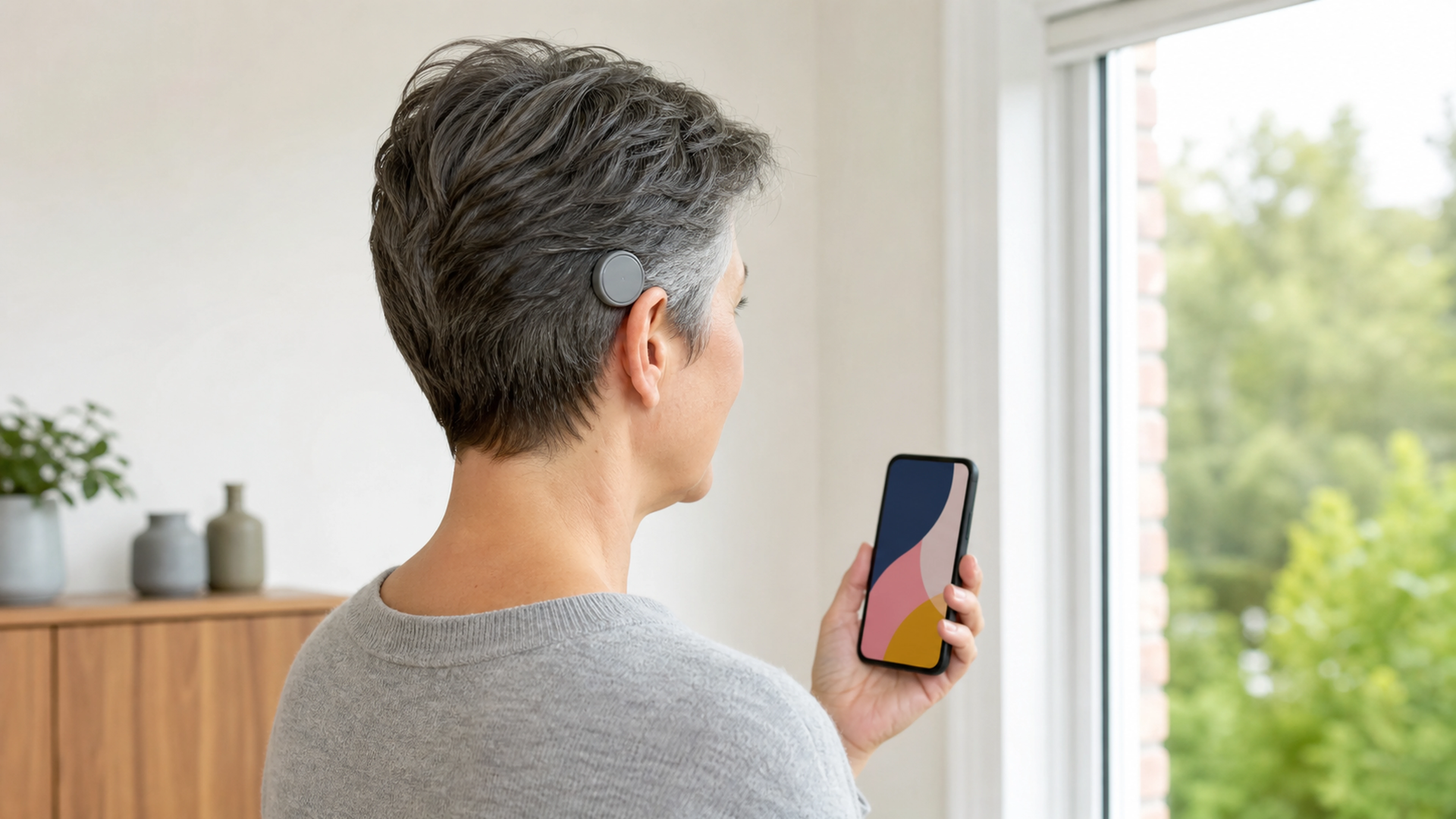 Personne portant un processeur auditif externe discret et tenant un smartphone près d’une fenêtre